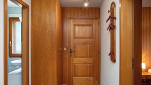 Eine Holztür mit Fußmatte am Eingang, ein kleines Regal mit Gegenständen und ein Blick in ein Badezimmer mit Waschbecken.