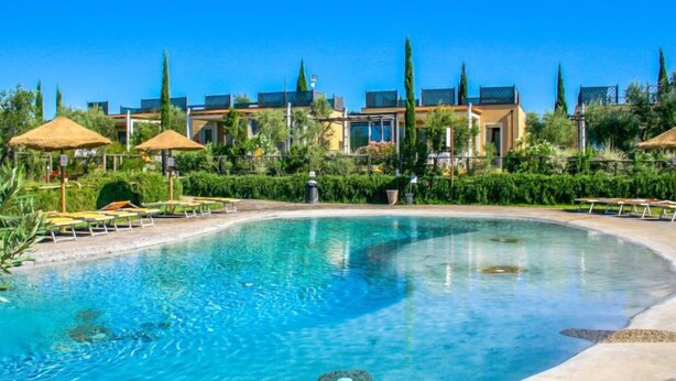 Una gran piscina nítida amb gandules i ombrelles, envoltada per vegetació ben cuidada i edificis moderns.