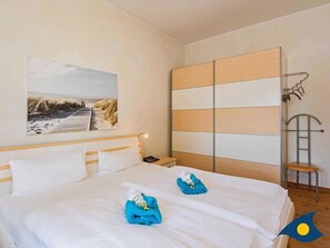 Una habitación de hotel con una cama grande, una mesita de noche, un televisor montado en la pared y un cuadro de una playa.