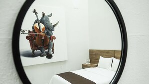 A bedroom with a bed, a nightstand, a mirror reflecting a painting of a rhinoceros playing a violin, and a wooden headboard. - Hotel Via Real (Atotonilco el Alto)