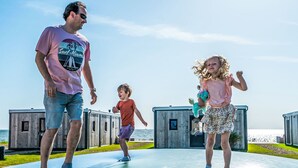 Uma família em piso de vidro transparente com vista para o oceano e estruturas de madeira ao fundo.