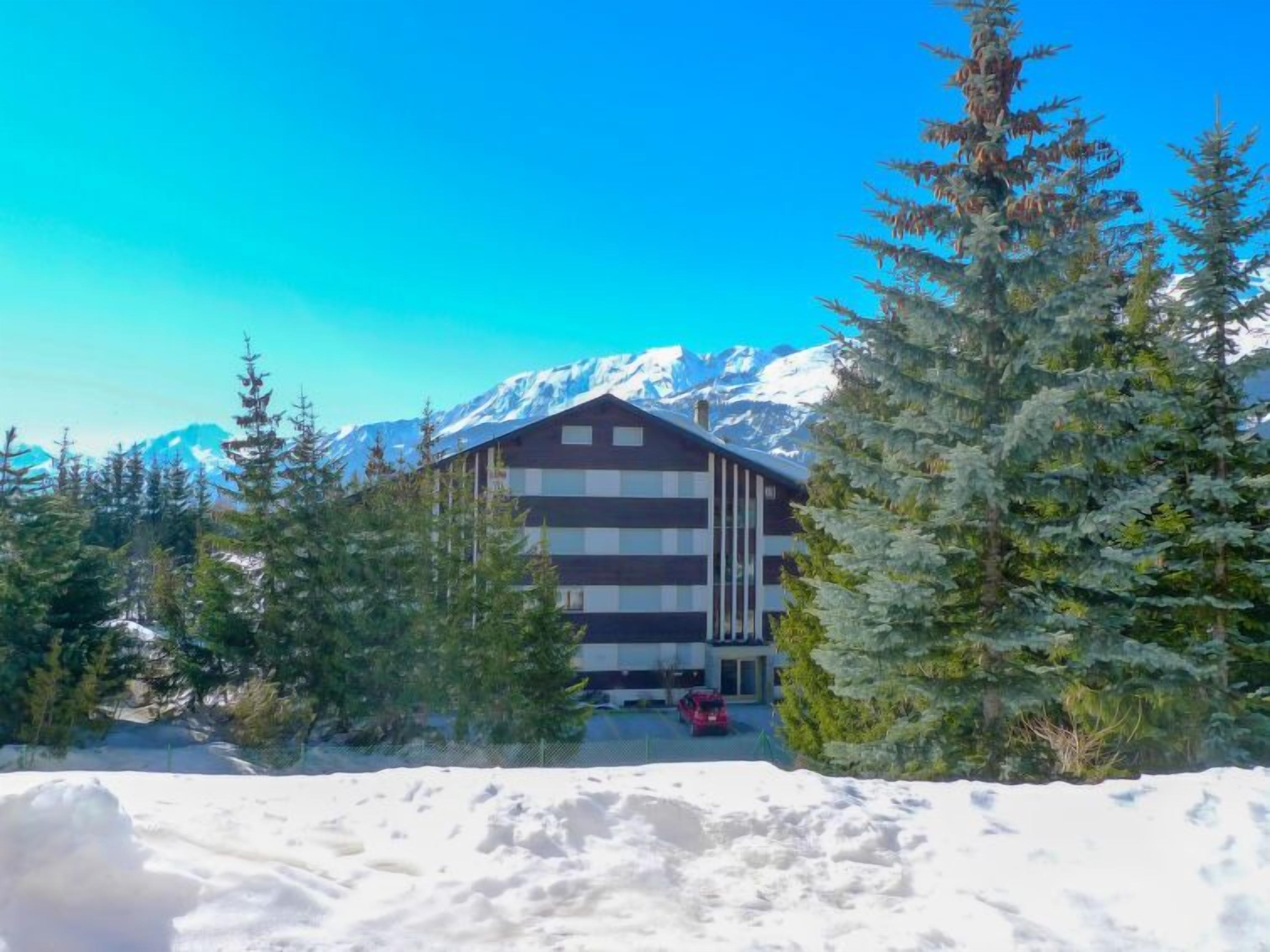 Un edificio con diseño moderno, rodeado de terreno nevado y árboles de hoja perenne, con una cordillera en el fondo.