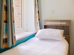 Een bed met witte lakens, een raam met gordijnen en een houten nachtkastje.