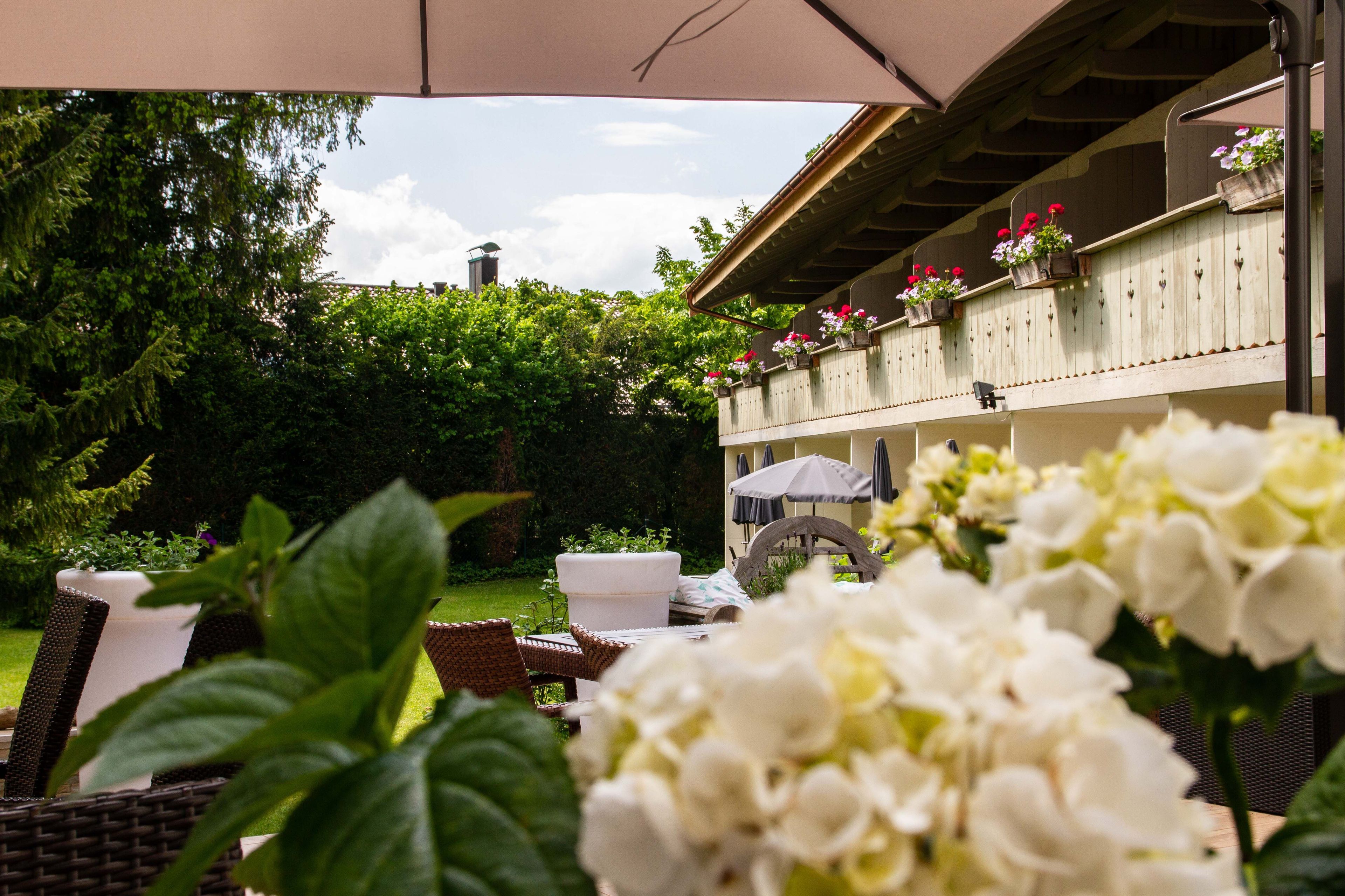 Un espacio al aire libre con mobiliario de mimbre, plantas en macetas y un edificio con jardineras florales al fondo.