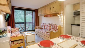 Ein gemütliches Zimmer mit einer Couch, einem Esstisch mit roten Tellern, einer kleinen Küchenzeile und einem großen Fenster.