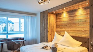 A hotel room with a wooden headboard, a bed with white linens, a desk with a chair, and a window with curtains. - Chesa Fleury by Interhome (St. Moritz)