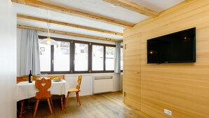 Una stanza con pavimento in legno, un tavolo da pranzo con sedie, una TV a parete e ampie finestre con tende.