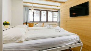 Una camera d'albergo con un letto, una televisione, una finestra con tende, un tavolo con una bottiglia e dei bicchieri, e una pianta.