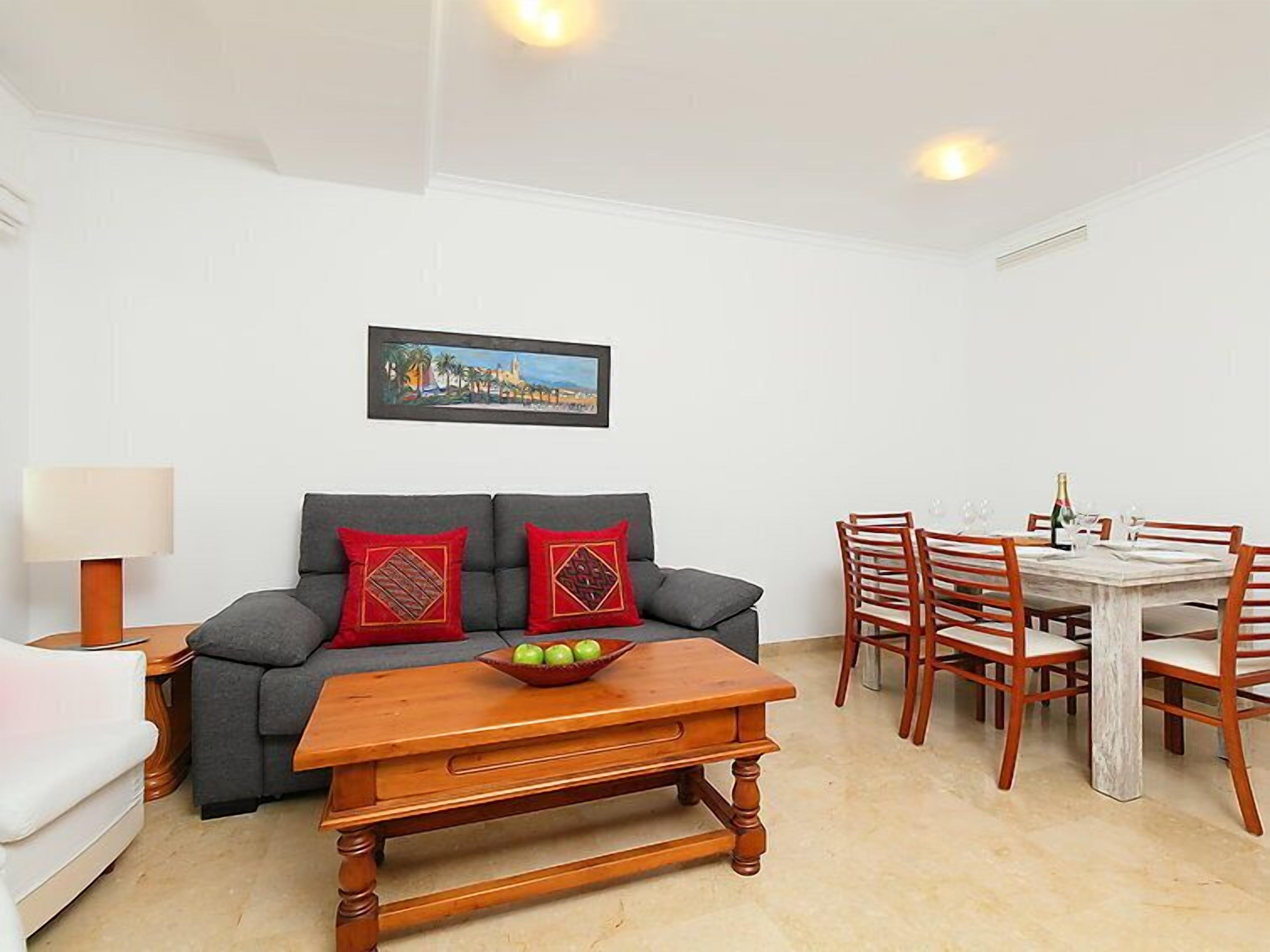 A living room with a grey sofa, red cushions, a wooden coffee table, and a dining area with wooden chairs and a table.