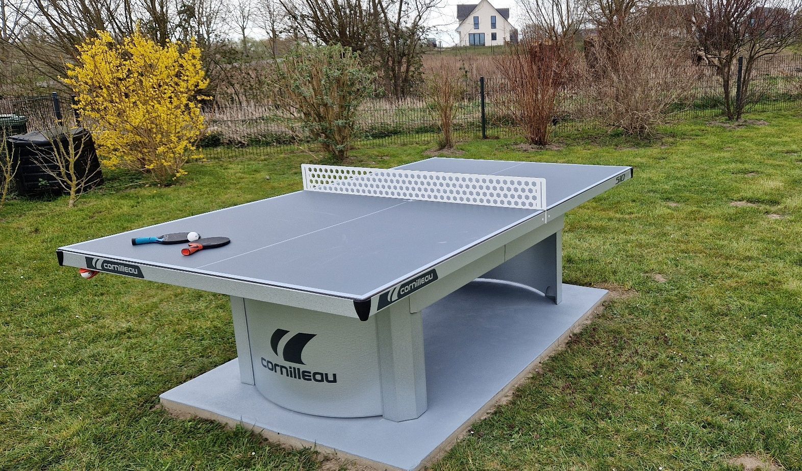 A Cornilleau ping pong table in an outdoor setting with a grassy lawn and trees in the background.