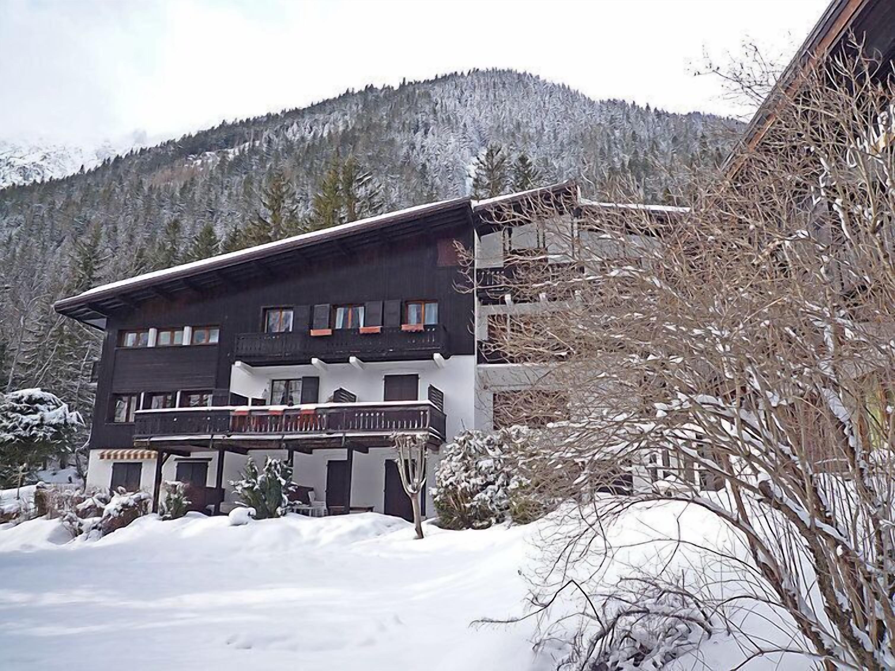 Een chalet met twee verdiepingen en een balkon, omgeven door een besneeuwde omgeving en een bebost berglandschap op de achtergrond.