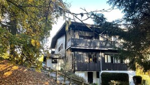 Une maison en bois à deux étages, dotée d’un balcon, entourée d’arbres et bordée d’un sentier recouvert de feuilles.