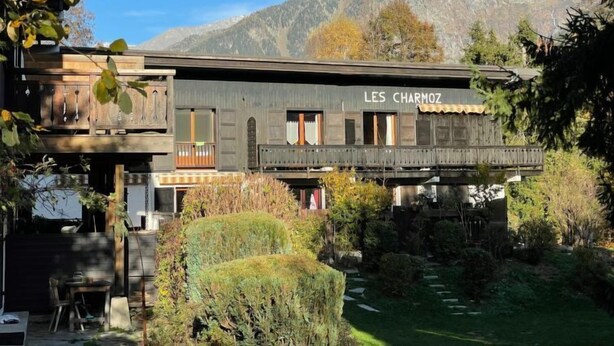 Un bâtiment rustique appelé « LES CHARMOZ », doté d’un balcon, entouré de verdure et avec des montagnes en toile de fond.