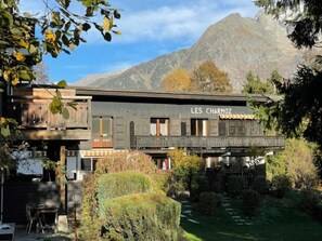 A rustic building named "LES CHARMOZ" with a balcony, surrounded by greenery and mountains in the background.