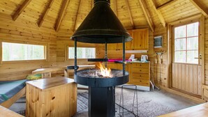A wooden cabin interior with a central wood-burning stove, wooden furniture, and a kitchen area.