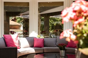 A curved sofa with cushions, a round table with a potted plant, and large windows with reflections of greenery and buildings. - Double Room 2 - Hotel Berlin Tegernsee Garni (Rottach-Egern)