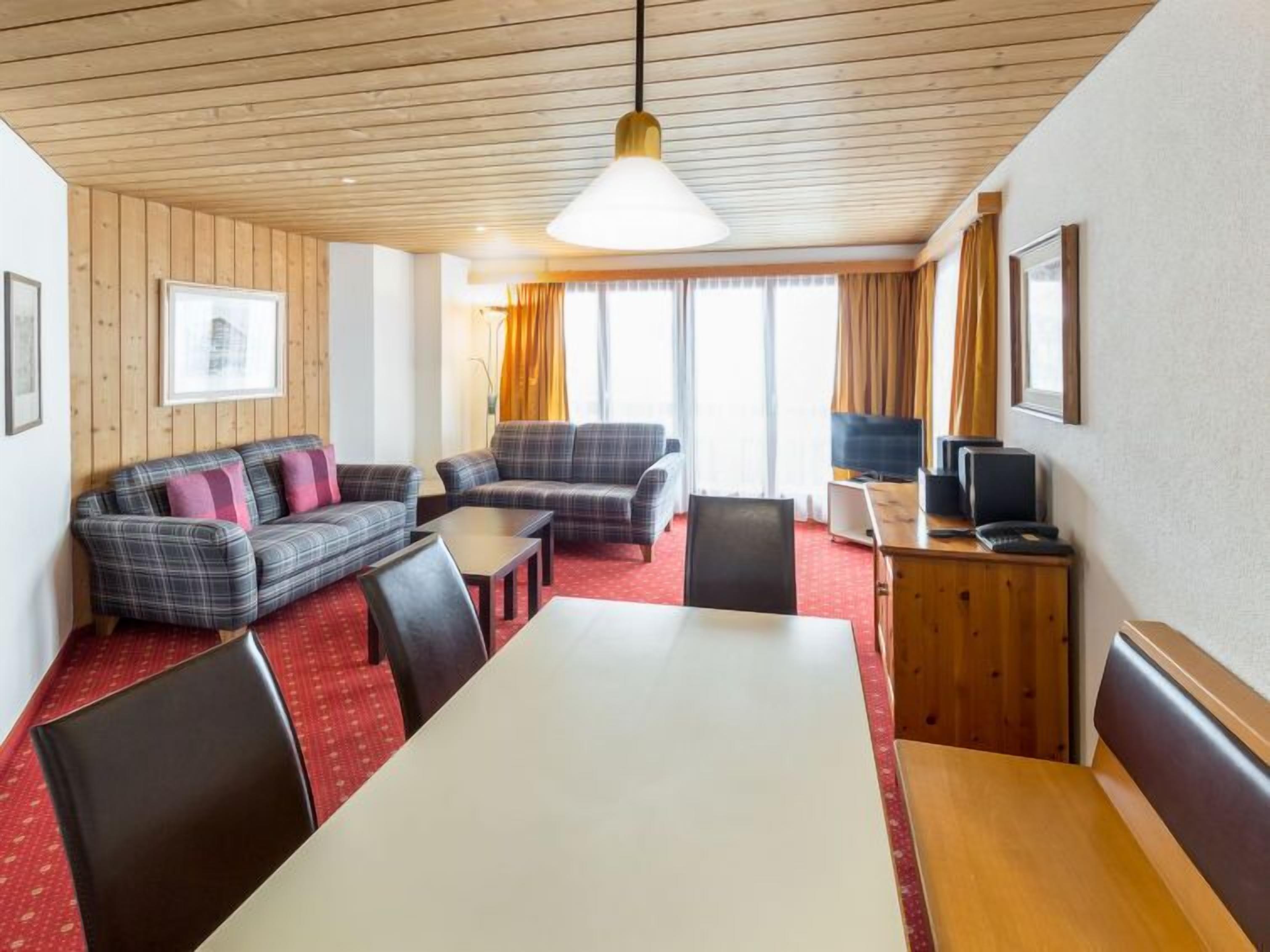 Una camera d'albergo con soffitto in legno, tappeto rosso, divano, tavolo da pranzo e televisore.