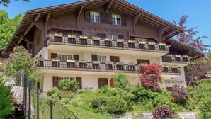 Un immeuble à plusieurs étages doté de balcons, entouré d’une végétation luxuriante et d’un mur de pierre.