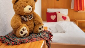 Un ours en peluche est assis sur une table en bois dans une pièce où se trouvent un lit, des coussins ornés de motifs en forme de cœur et une lampe fixée au mur.