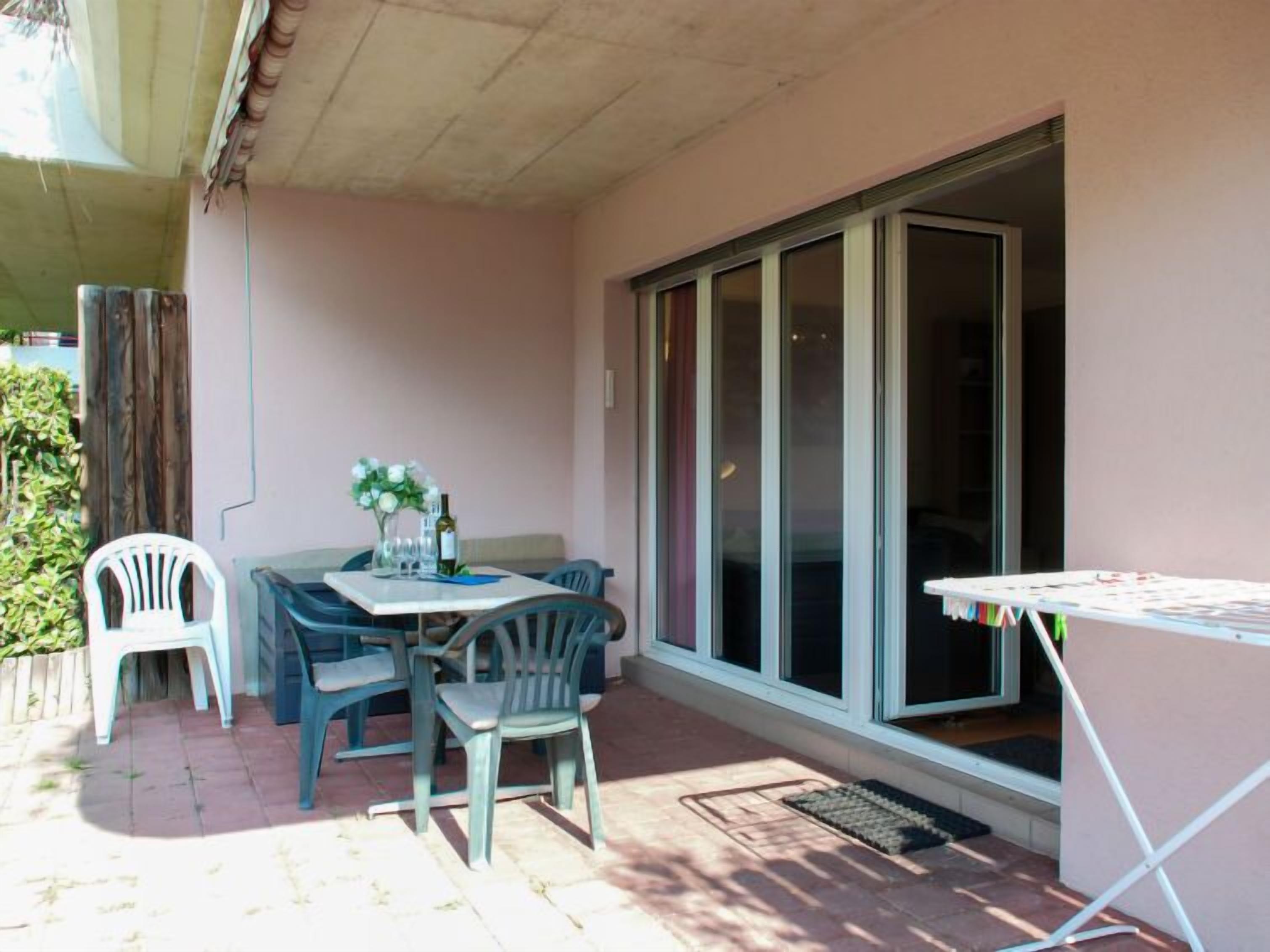 Uma área de pátio com mesa e cadeiras, uma pequena mesa com uma garrafa e copos, e uma porta de vidro deslizante.