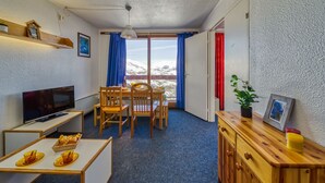 A room with a TV, wooden furniture, a dining table, and a view of snow-capped mountains through large windows. - Lunik Orion by Interhome (Le Corbier)