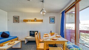 A cozy room with a wooden dining table set for four, a blue sofa, a small TV, and a window with a view of a snowy landscape. - Lunik Orion by Interhome (Le Corbier)