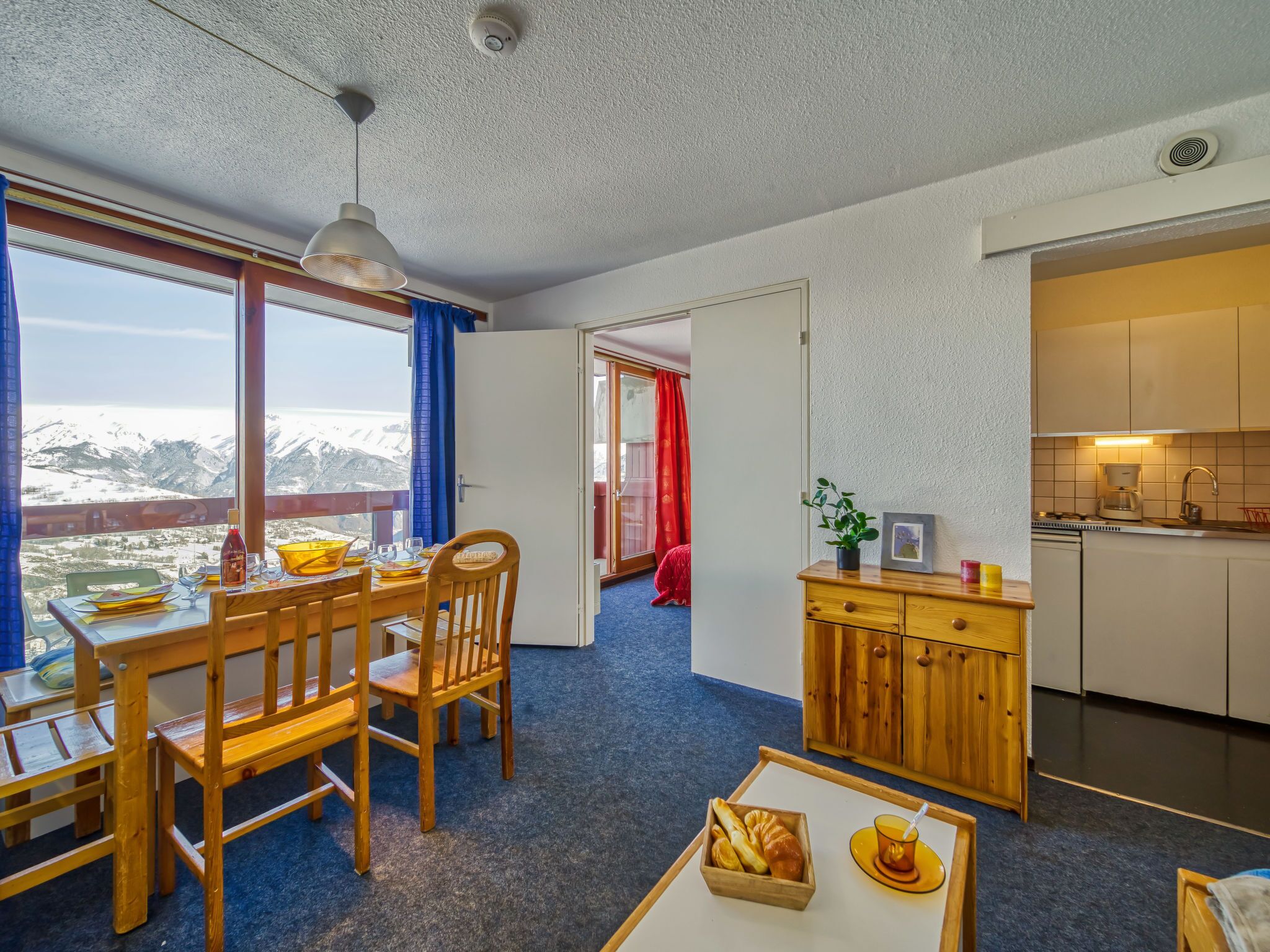 Una habitación acogedora con una mesa de comedor, sillas, una pequeña cocina y vistas a montañas nevadas.