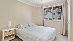 Une chambre à coucher avec un lit, une table de chevet avec une lampe, une fenêtre avec des rideaux et une vue sur un bâtiment à l’extérieur.
