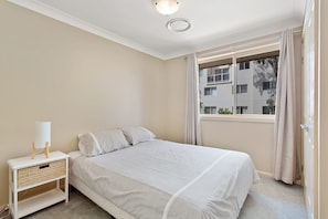A bedroom with a bed, a bedside table with a lamp, a window with curtains, and a view of a building outside. - Ajax Avenue, 6 (Nelson Bay)