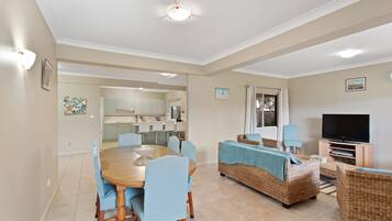 A spacious living area with a dining table, chairs, a television, and a kitchen in the background.