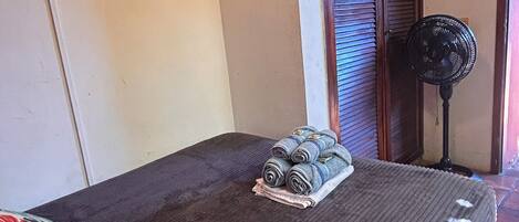 A bedroom with a bed, a pillow, a rolled-up towel, a standing fan, and a wall socket.