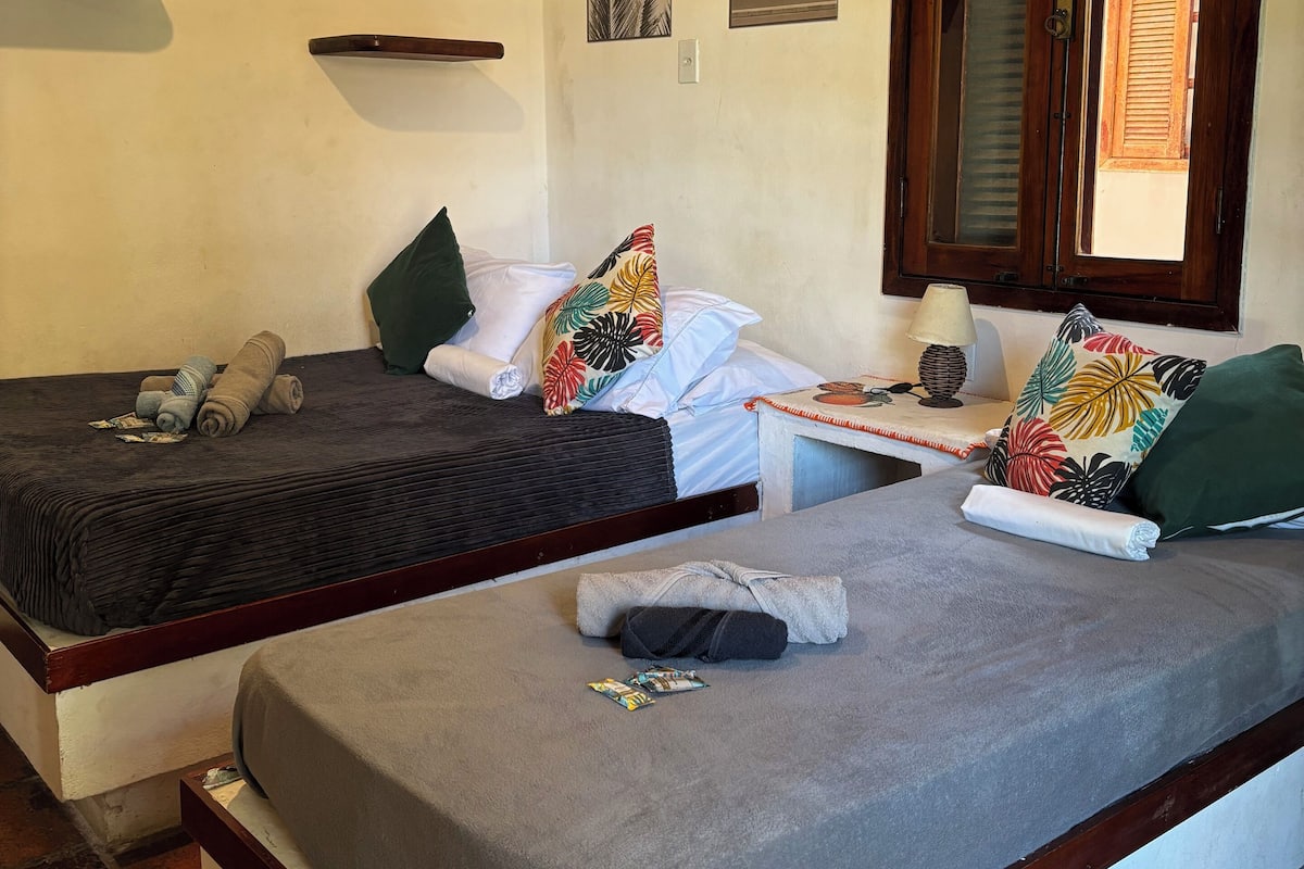 A room with two single beds, a small table with a lamp, and a window with wooden shutters.
