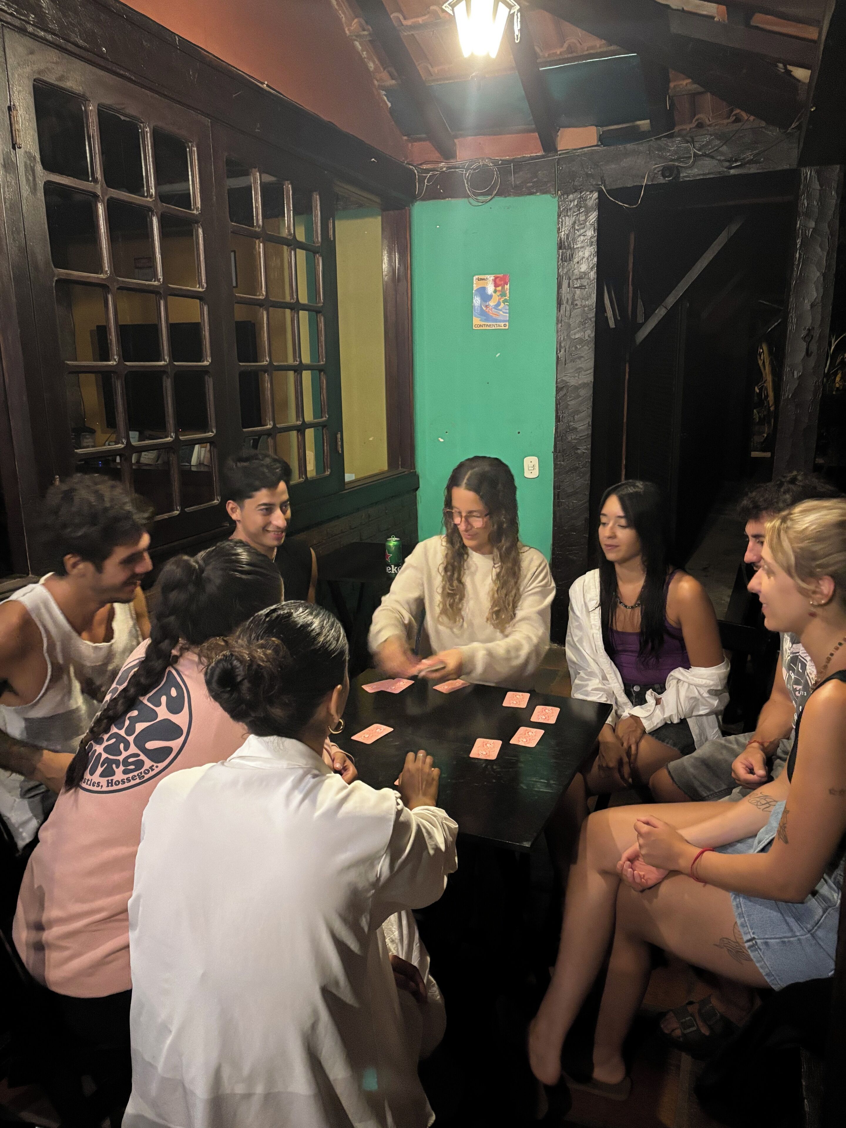 A group of people playing cards around a table in a room with wooden walls and a green door.