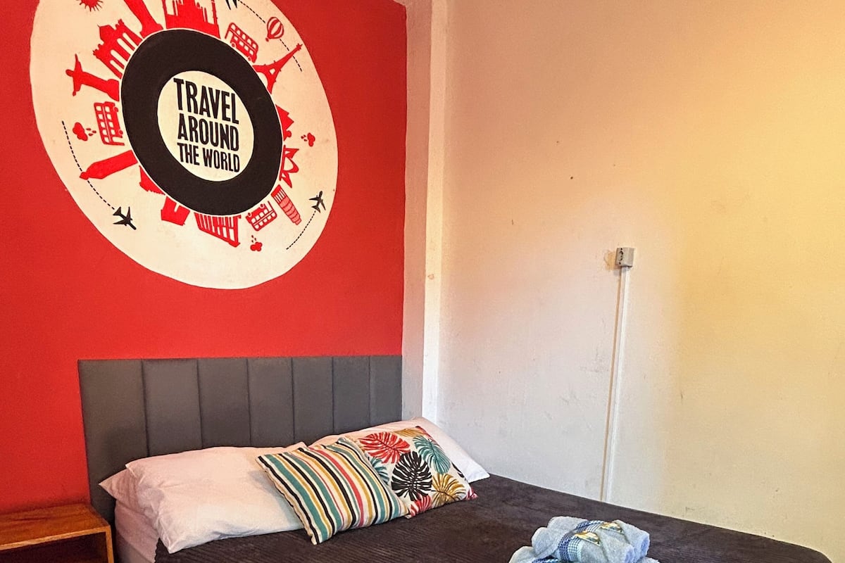 A hotel room with a red wall, a bed with a dark headboard, a pillow with a colorful pattern, and a tray with towels.