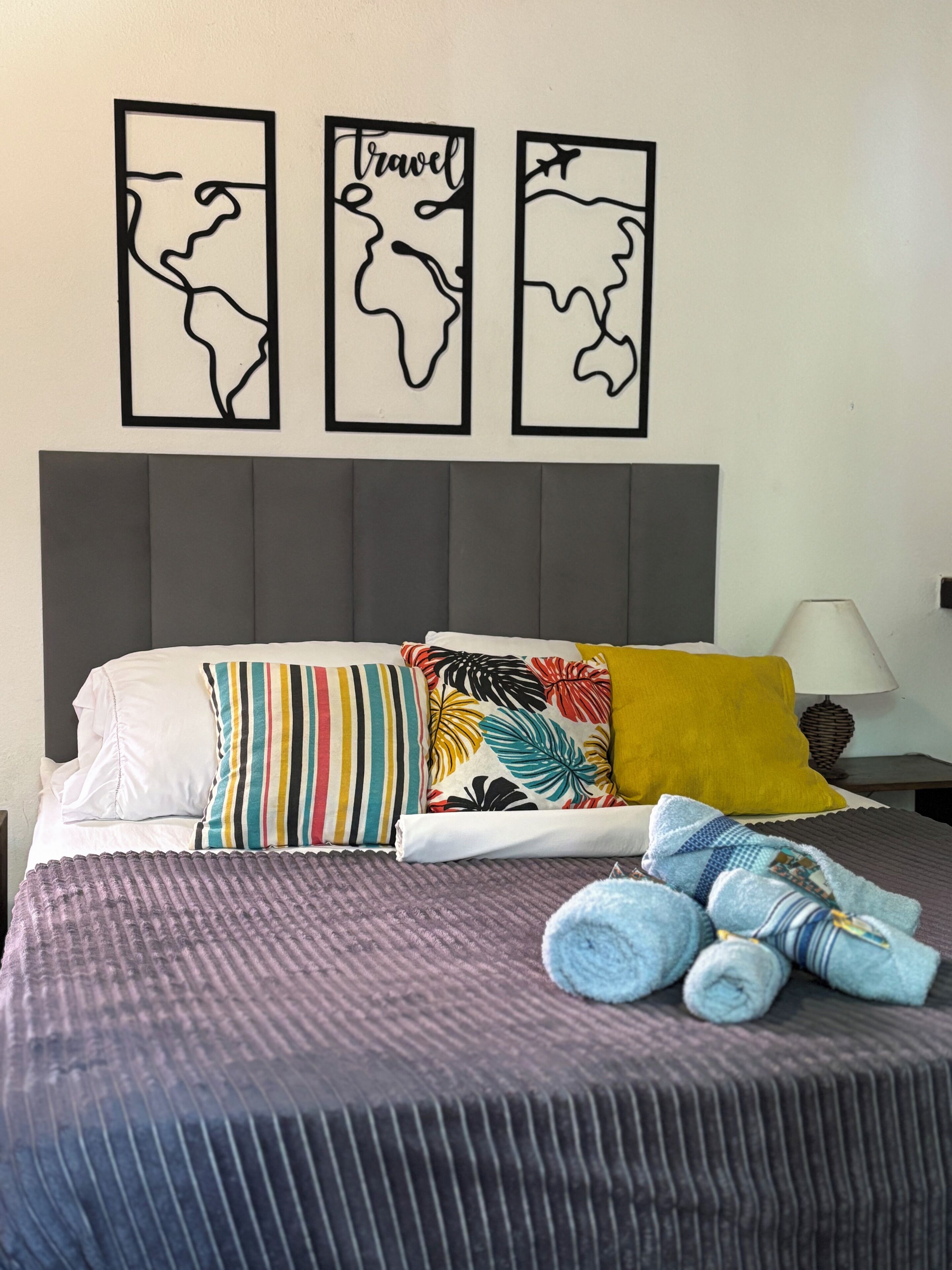 A neatly made bed with striped and tropical patterned pillows, a grey headboard, and three wall art pieces depicting abstract maps.