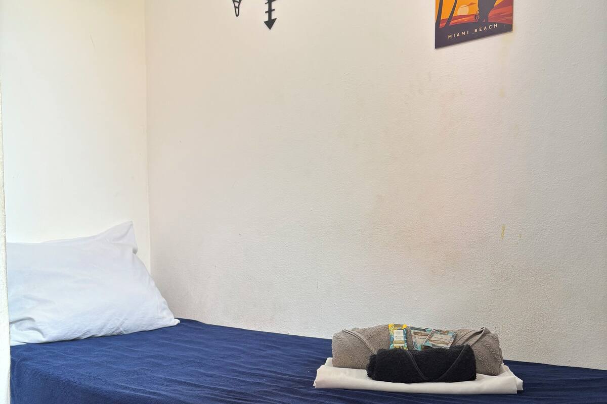 A single bed with a blue mattress and a white pillow, a small decorative wall piece, and a poster of a sunset over Miami Beach.