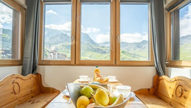 A dining area with a wooden table and benches, a bowl of fruit, and a view of mountains through large windows. - Arcelle 505 by Interhome (Val Thorens)