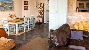 Un salon chaleureux avec un canapé en cuir marron, un coin repas avec une table et des chaises en bois, et une cuisine avec un mur en pierre et un meuble blanc.