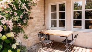 Une terrasse avec une table et des chaises en bois, un mur en pierre et une fenêtre donnant sur un paysage verdoyant.