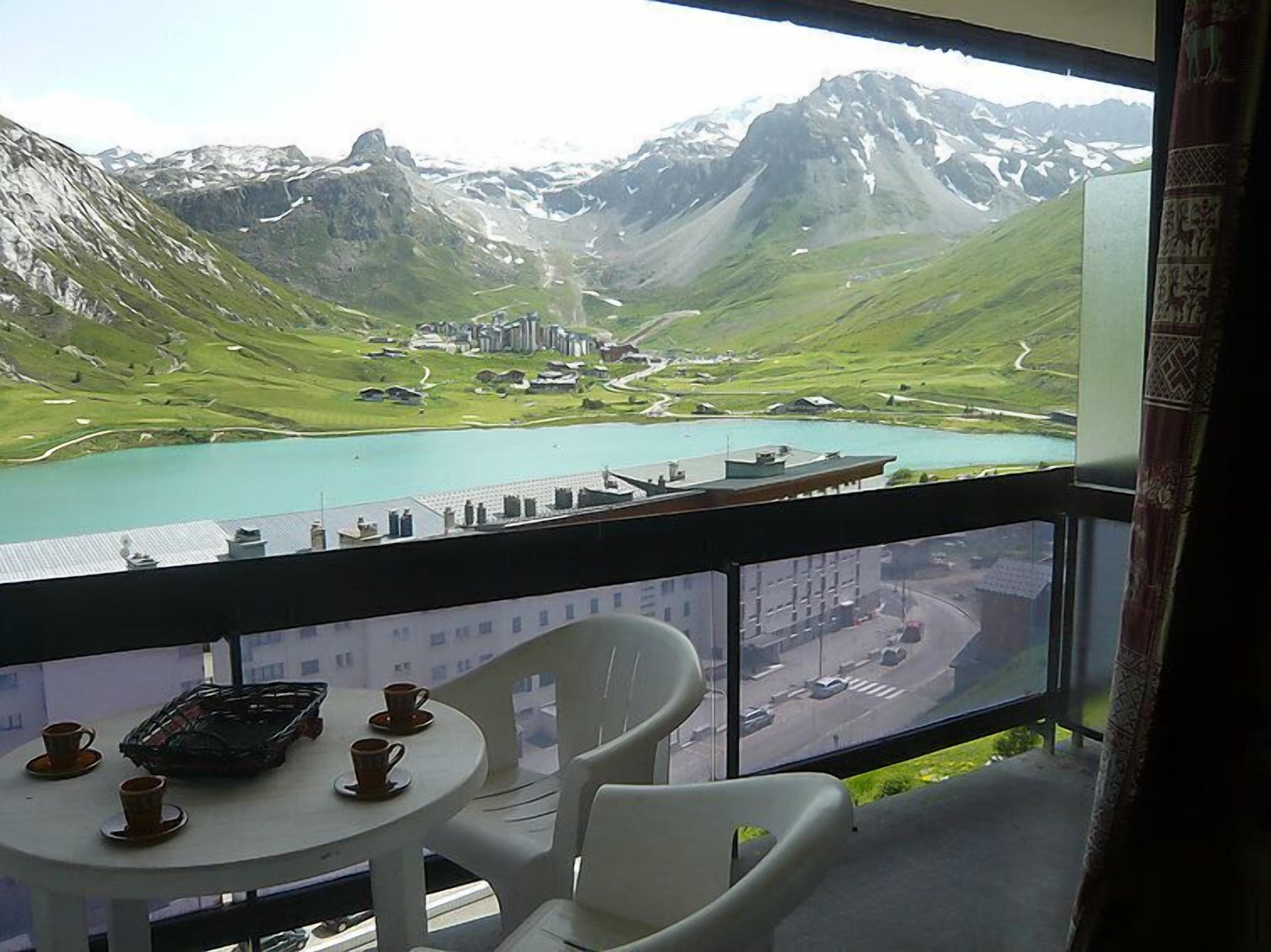Een balkon met een tafel en stoelen, met uitzicht op een bergachtig landschap met een meer en gebouwen.