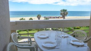 A balcony with a table set for two, overlooking a beach with palm trees and a clear blue sky.