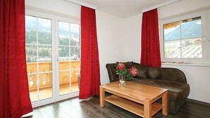 A living room with a brown sofa, a wooden coffee table, and a vase with flowers. There are red curtains and a view of a balcony with a wooden railing. - Pfeifer by Interhome (Pians)