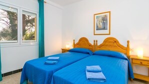 Habitación doble con dos camas individuales, cabecera de madera, mesita de noche con lámpara y ventana con cortinas.