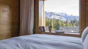Un dormitorio con un ventanal que ofrece vistas a la montaña, una cama con ropa blanca, una cortina y una pequeña mesa con libros y una planta.