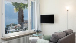 Een woonkamer met een groot raam, een flatscreen televisie aan de muur, een radiator onder het raam en een bank met kussens.