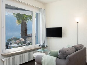 A living room with a large window, a flat-screen TV mounted on the wall, a radiator below the window, and a sofa with cushions.