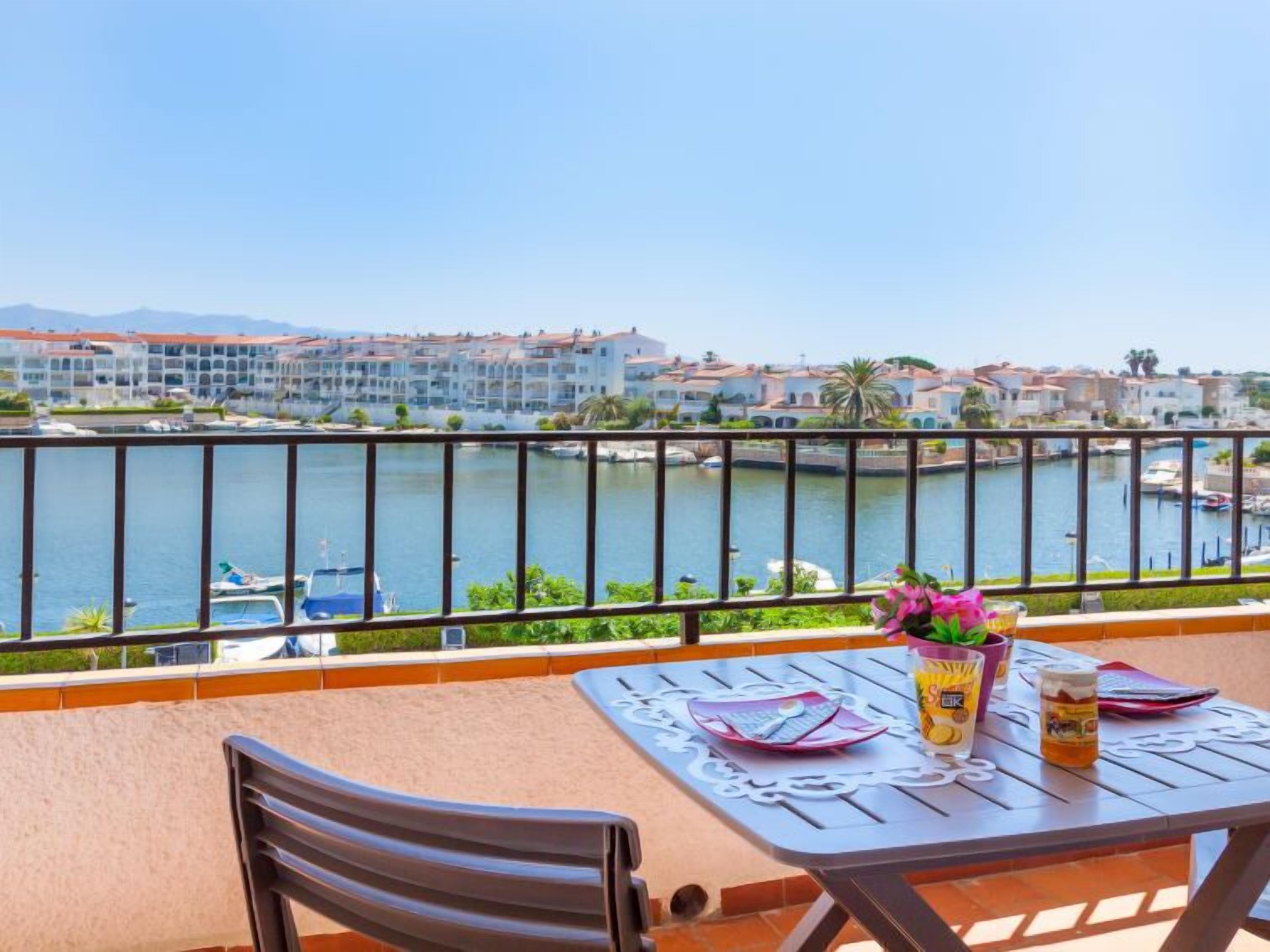 Een balkon met een tafel gedekt voor twee personen, met uitzicht op een jachthaven met boten en gebouwen op de achtergrond.