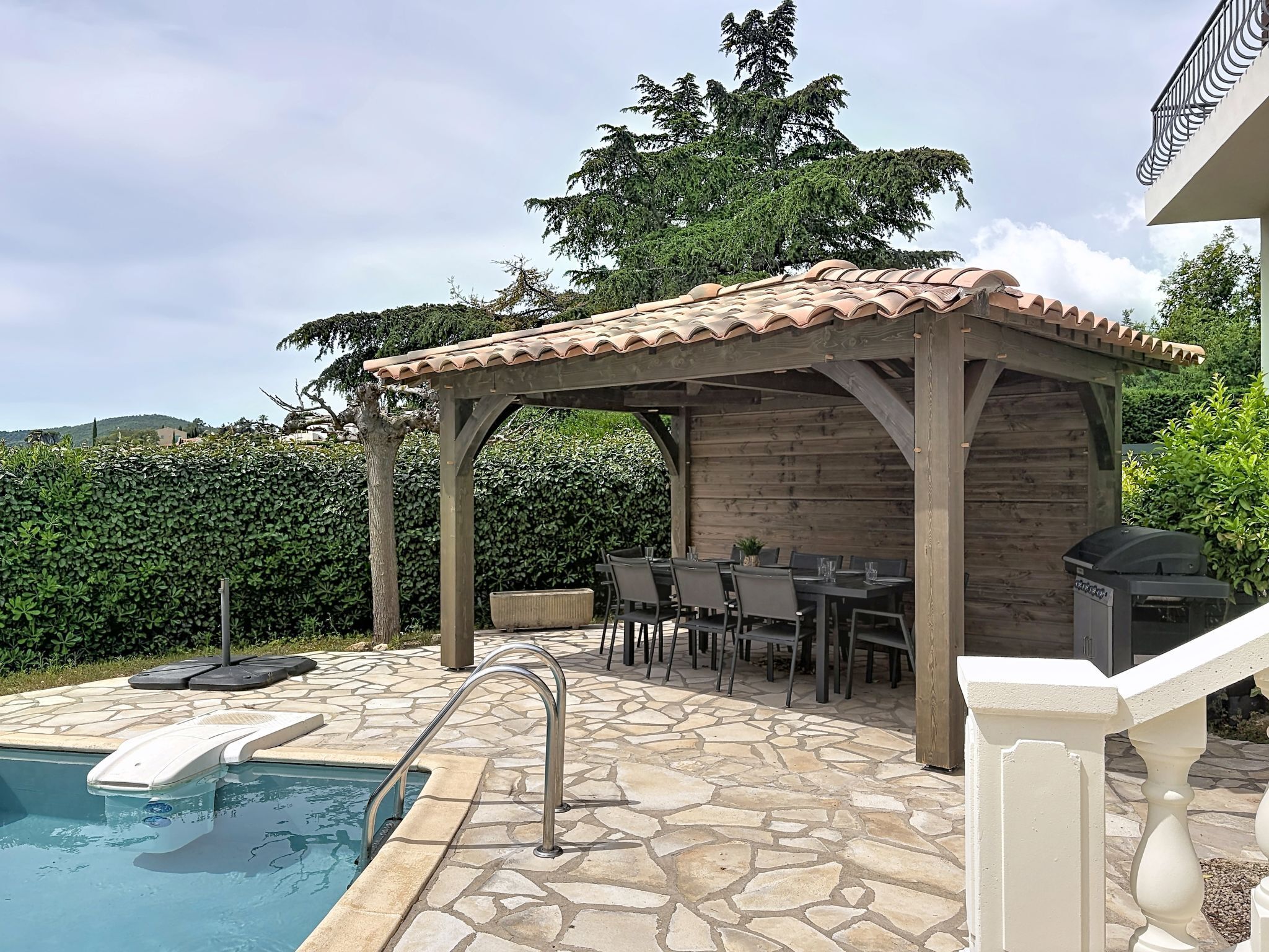A pool with a stone deck, a covered patio with tables and chairs, and a pergola with a tiled roof.