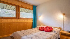 A cozy room with a wooden paneled wall, a bed with a beige comforter, two red pillows, a nightstand with a lamp, and a window with a view of a snowy landscape. - Brelin Danchet 1026 by Interhome (Les Menuires)