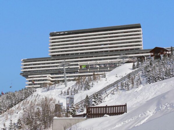 Un bâtiment moderne à plusieurs étages, avec un télésiège visible en arrière-plan, entouré d’arbres couverts de neige et d’une piste de ski.